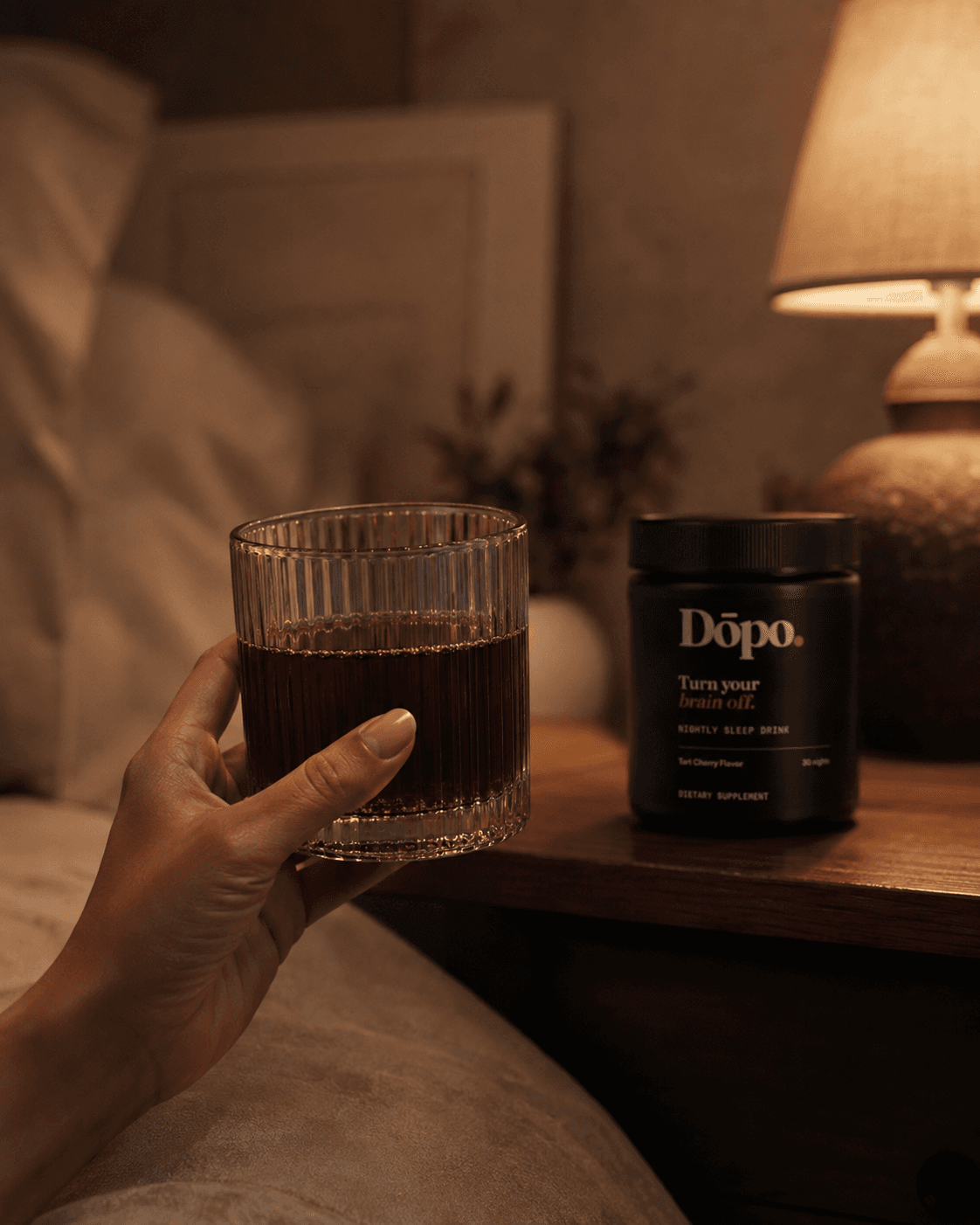 A hand lifting a glass of ruby Dōpo in warm bedside light, the tub soft-focus behind it