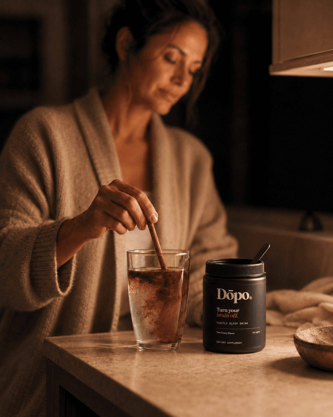 A person stirring a ruby Dōpo drink in a warm kitchen at night, with the Dōpo tub clearly visible beside the glass